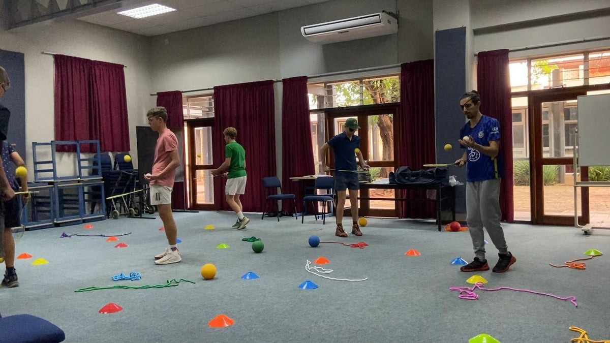 A wide shot of a hall drill, teenagers and two coaches among cones and balls