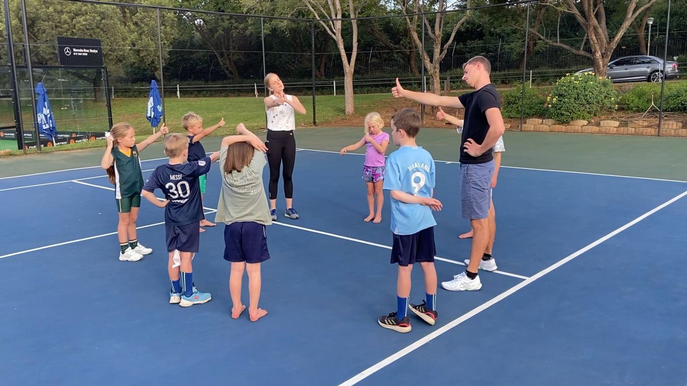 Jannik and Maria with a circle of children on the blue court, thumbs up